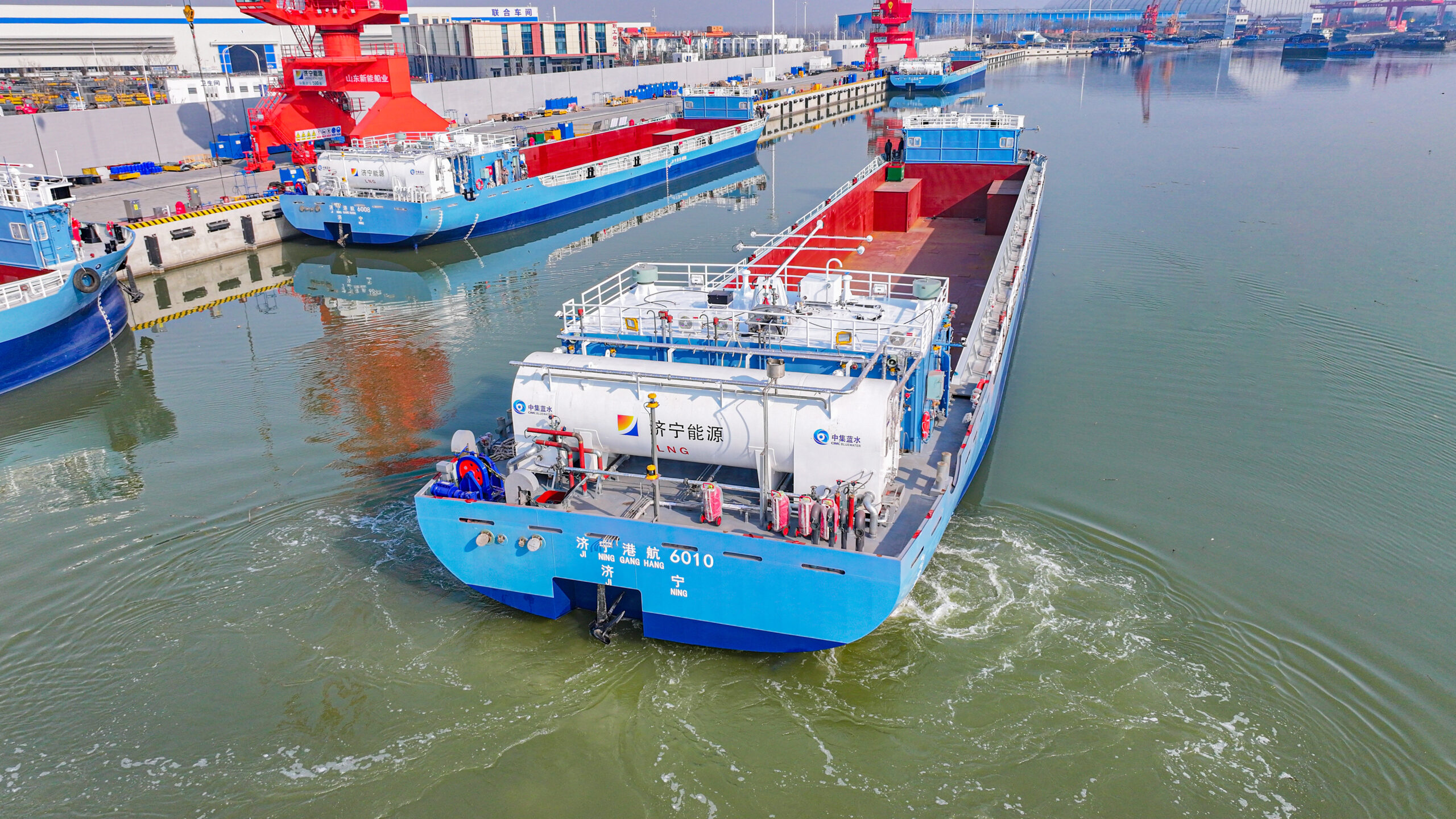 Aerial view of a blue chemical tanker ship with white cylindrical tanks navigating a busy port.