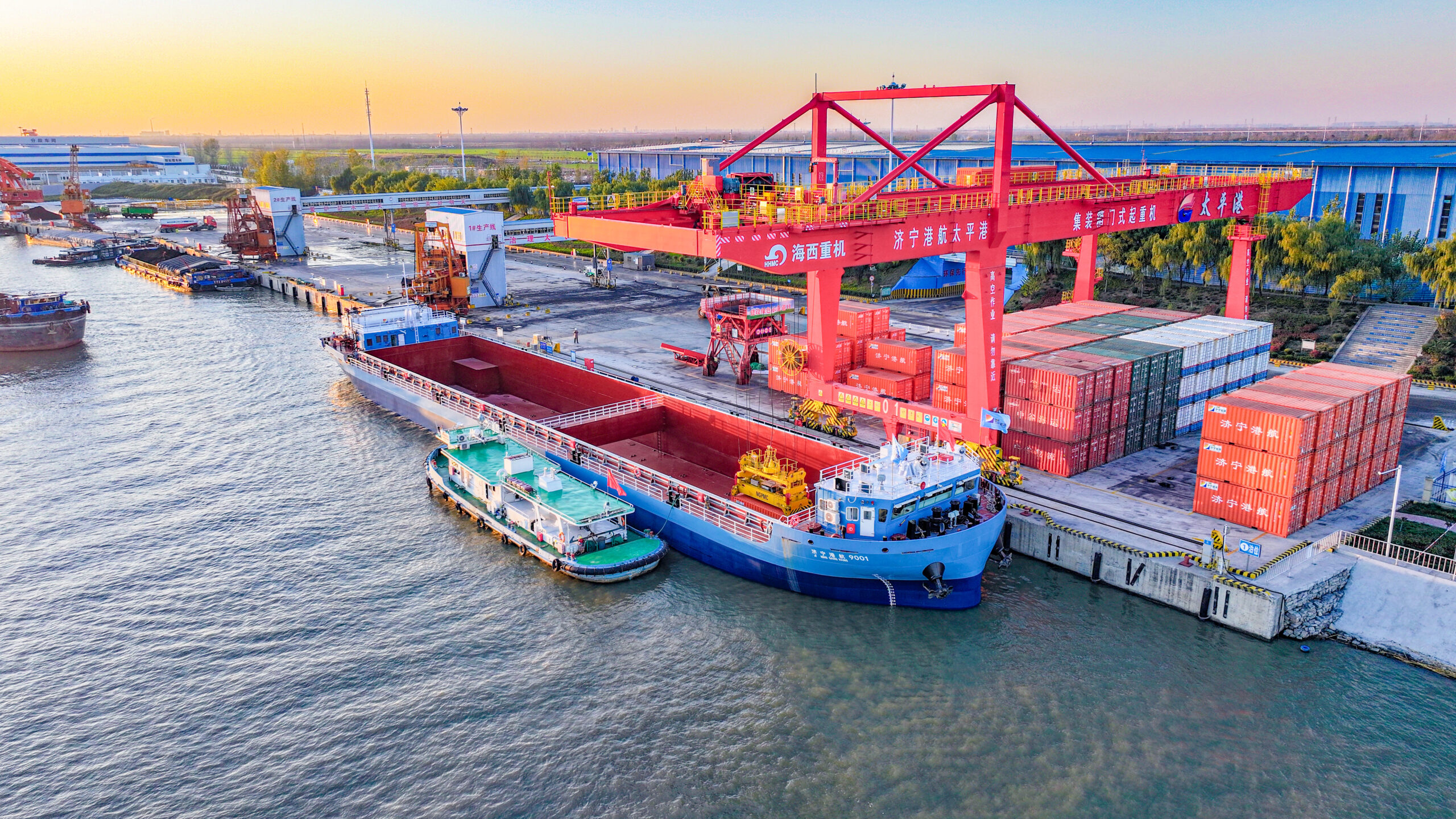 Aerial view of a busy port at sunset with red gantry cranes loading a bulk carrier ship and stacked containers along the waterfront.