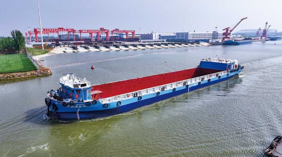 A blue cargo barge loaded with red material navigates a river beside red gantry cranes and port facilities.