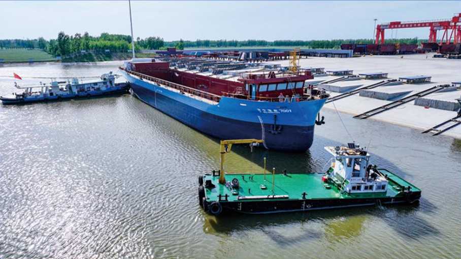 A large blue cargo ship docked at a river port, assisted by tugboats and a green barge amid cranes and warehouses.