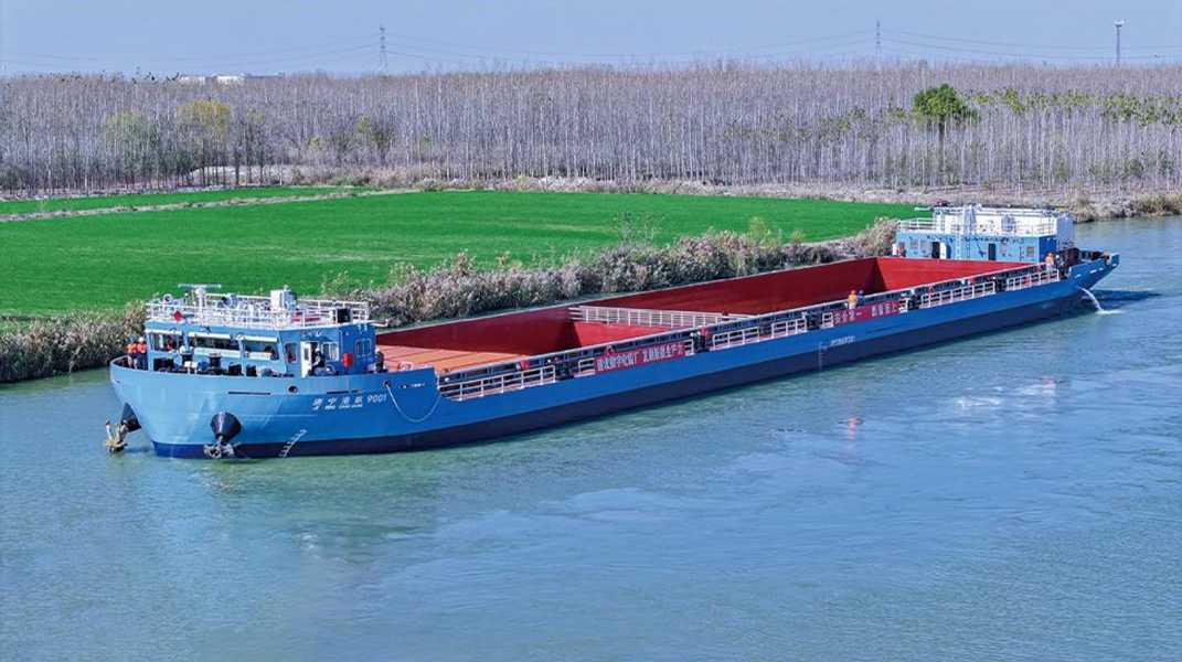 90m A blue cargo barge loaded with red bulk material sails along a river bordered by green fields and bare trees.