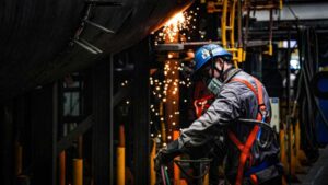 worker in protective gear and harnesses performs welding on a large metal structure in an industrial setting, with sparks flying.