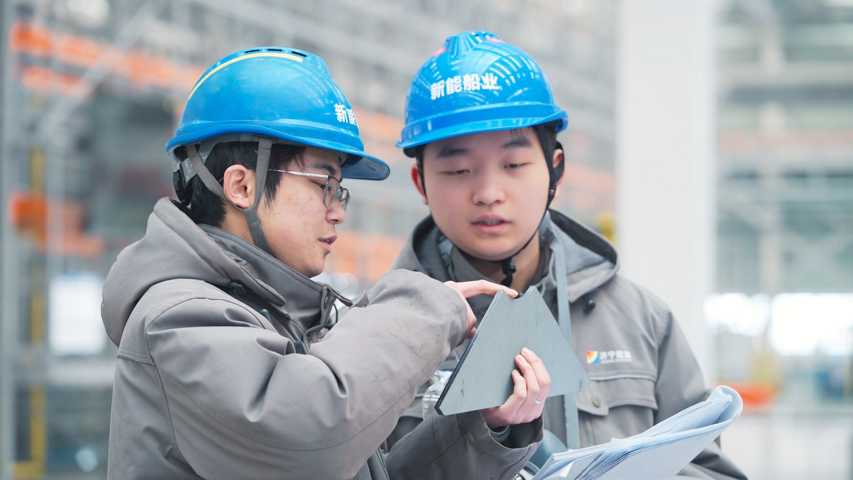 Two workers in blue hard hats and gray jackets discuss a triangular metal panel in a factory setting.