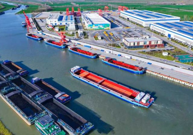 Aerial view of a busy river port with docked cargo ships, red cranes, and industrial warehouses along the waterway.