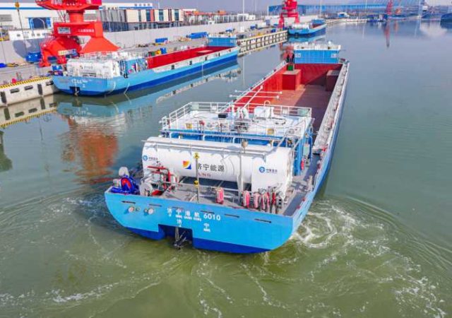 Aerial view of a blue vessel with an LNG tank on the rear navigating a busy port with red cranes.